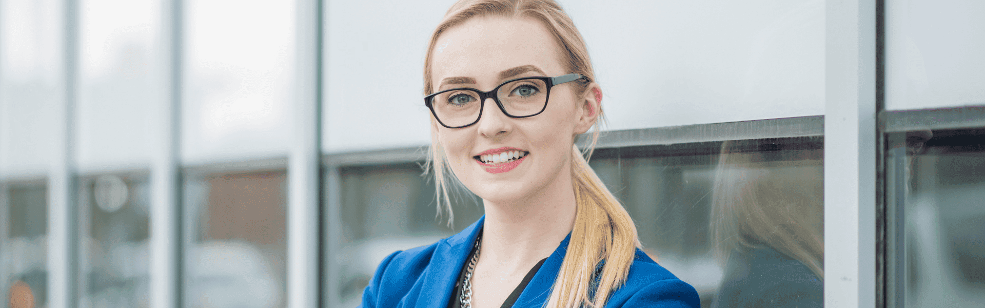 junge Frau mit blonden Haaren lächelt in die Kamera und steht vor einem Bürogebäude